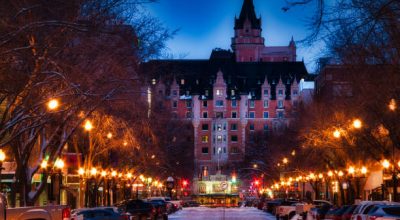 A snow cover street illuminated by street lights on both sides with a tradition Canadian style building at the end of the street with a glowing blue sky.